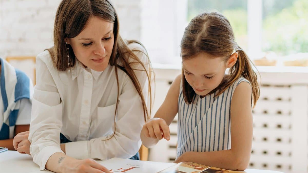 A teacher guides a young girl reading a book indoors, emphasizing learning and connection.