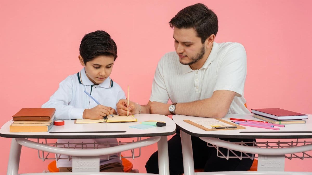 A teacher helps a student with schoolwork at a desk against a pink background.