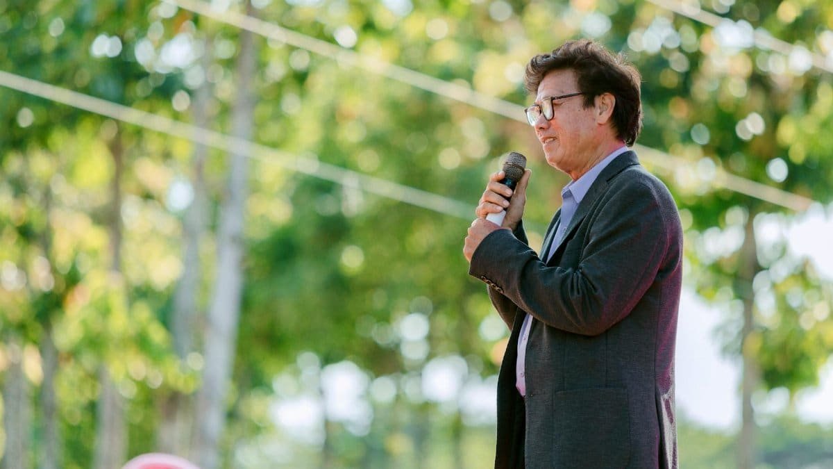 A confident man speaking outdoors, surrounded by trees, holding a microphone.