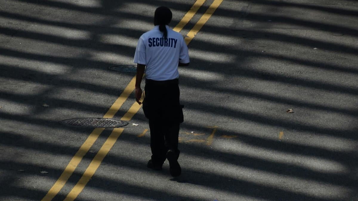 A security guard walks down a sunlit street, casting shadows on the pavement.