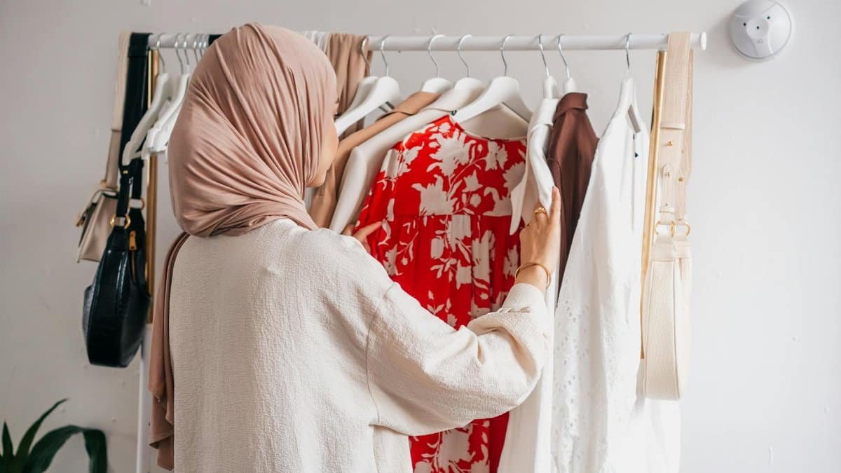 Woman in hijab organizing wardrobe, selecting various clothing options indoors.