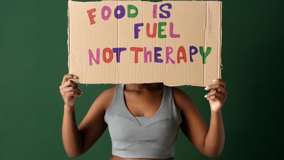 A woman holds a sign saying 'Food Is Fuel Not Therapy' against a green background.