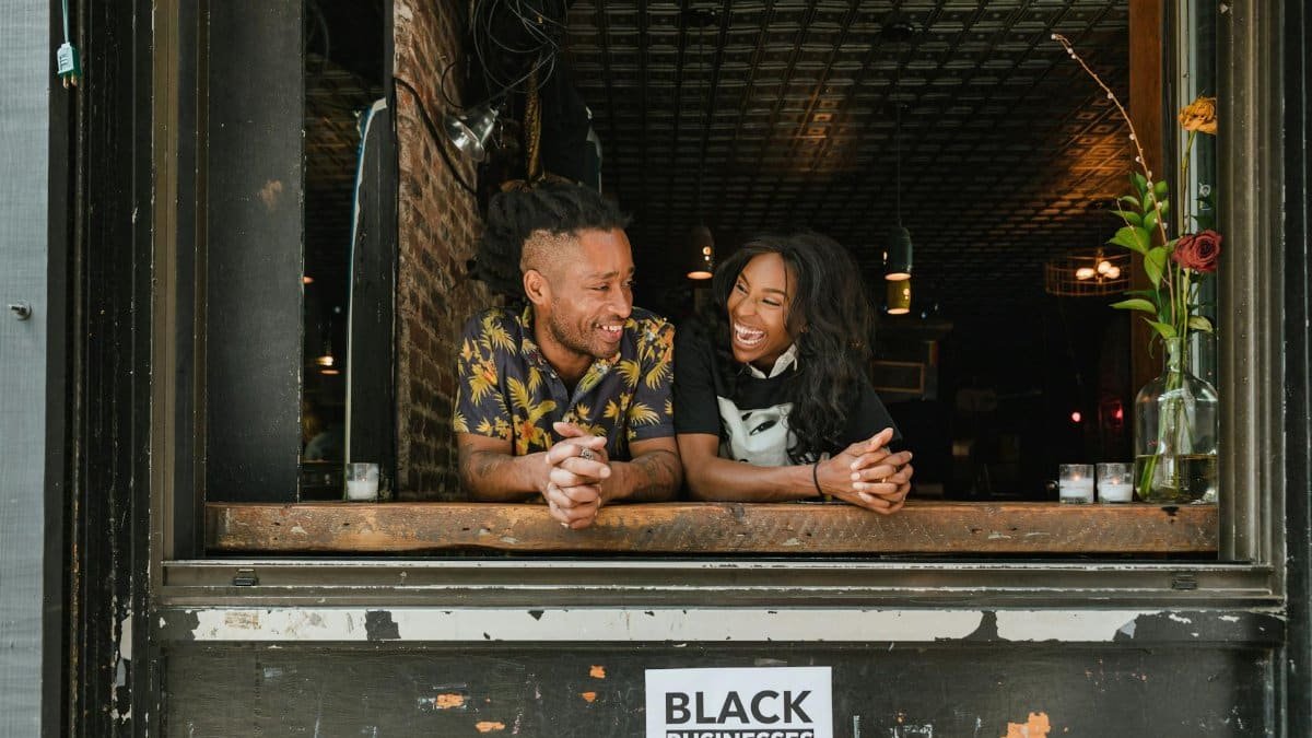 Two joyful business owners lean on a window sill with a 'Black Businesses Matter' sign visible.