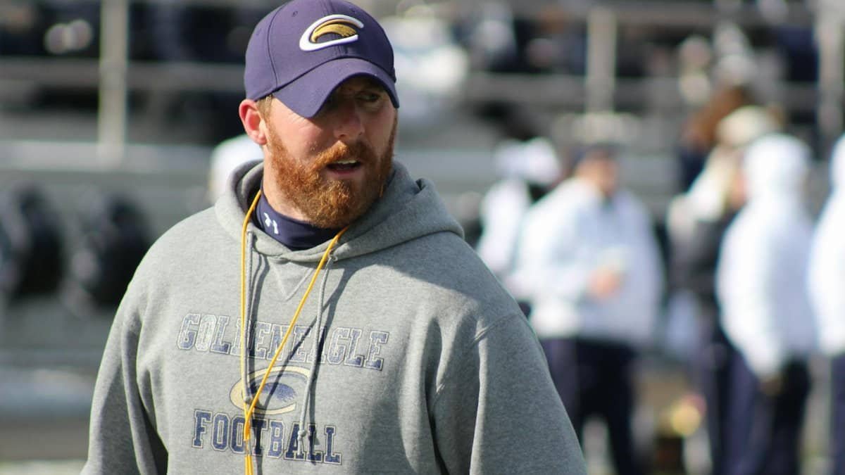 A focused football coach in a hoodie and cap on the sidelines of a stadium, ready to lead the team.