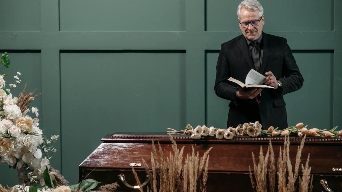 Pastor conducting a funeral service, standing beside a coffin adorned with flowers. Indoor setting.