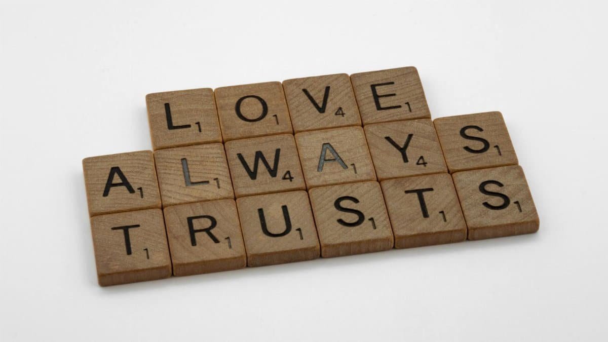 Close-up of wooden Scrabble tiles spelling 'Love Always Trusts' on white surface.