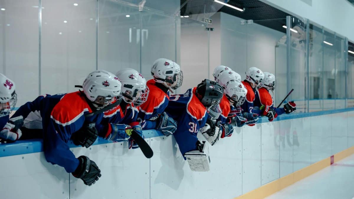 Group of young hockey players in full gear leaning on the railing in an indoor ice rink.
