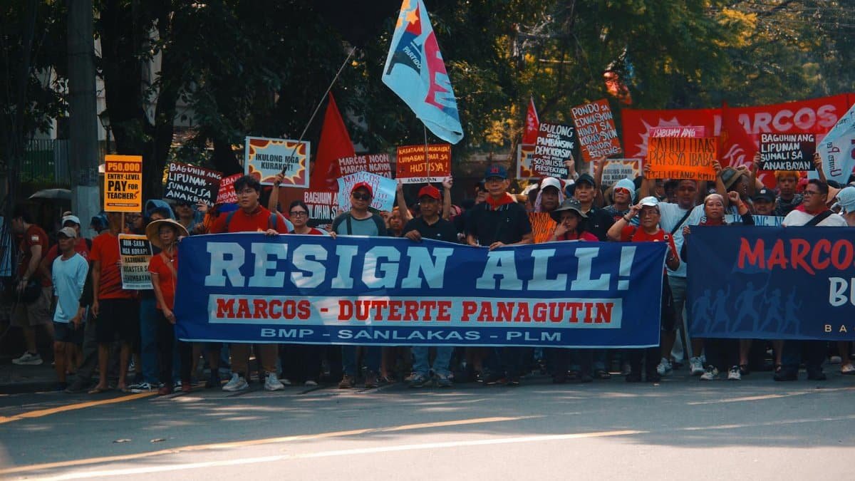 Crowd gathers in street protest holding signs demanding resignation with political messages.