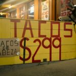 Street view of a taco stand with large yellow sign advertising tacos for $2.99, captured at night.