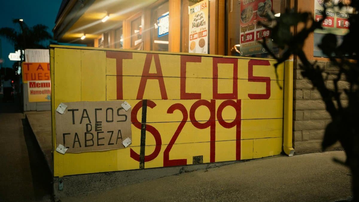 Street view of a taco stand with large yellow sign advertising tacos for $2.99, captured at night.
