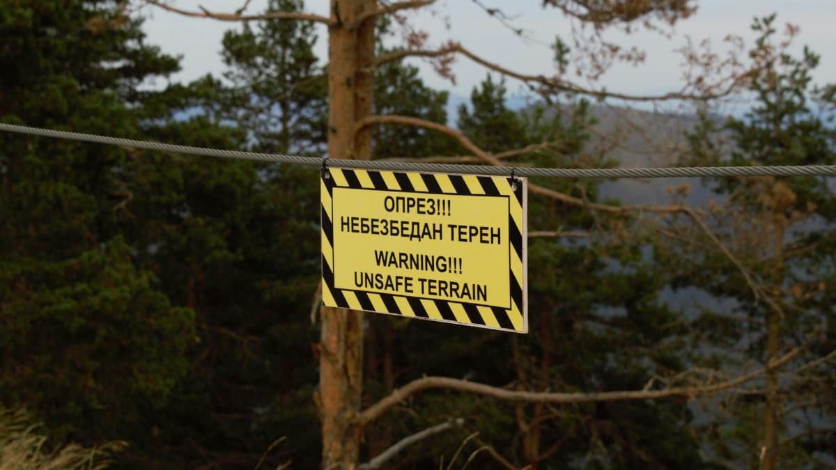 A warning sign cautions about unsafe terrain in a forested area, indicating potential danger.