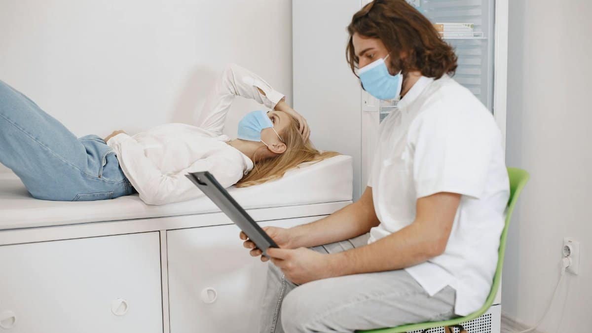 Doctor consulting patient wearing face masks in a medical examination room, discussing healthcare plan.