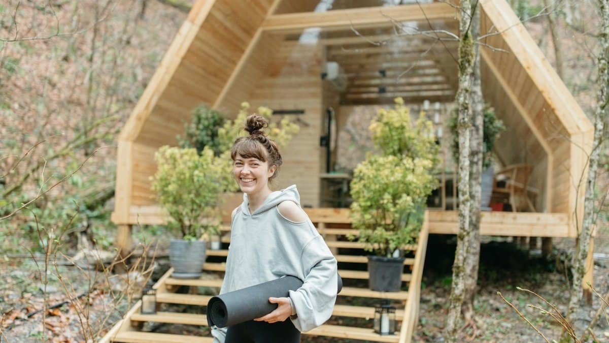 Cheerful woman with yoga mat in front of a wooden cabin surrounded by nature.