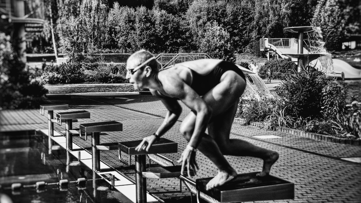 Black and white side view full body sporty swimmer in swimming suit and goggles standing on block in track start position preparing to dive in outside pool