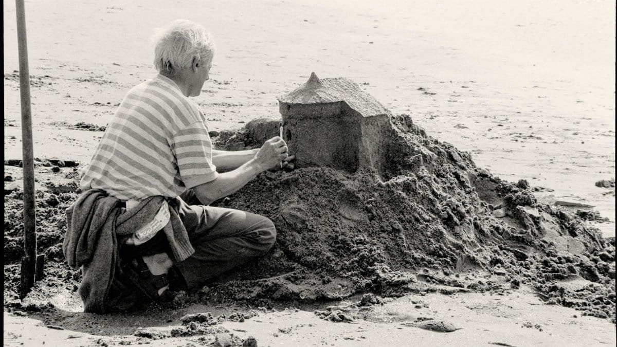 A man diligently builds a sandcastle on a serene beach in black and white.