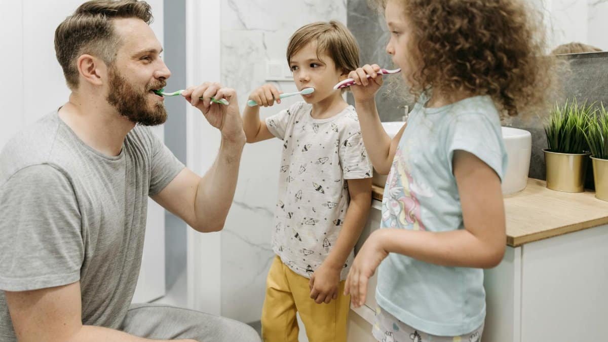 A father and his two children brushing their teeth in a bright bathroom.