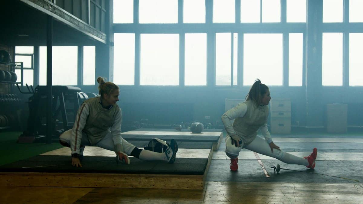 Two female fencers in gear stretching indoors, preparing for exercise.