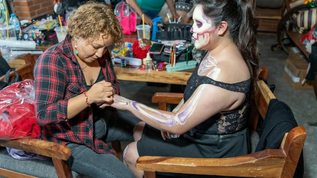 Two women preparing for Día de los Muertos with intricate face and body art in Mexico City.