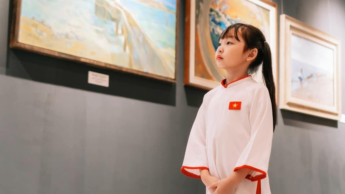 A young girl in traditional Vietnamese attire observes paintings in an art gallery.