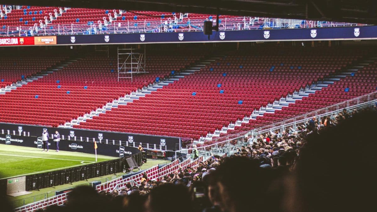 Panoramic view of stadium with empty red seats and crowd gathered for a live match.
