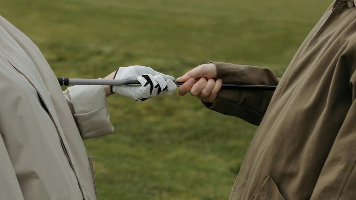 Two golfers in coats share a golf club on a grassy course, highlighting leisure and companionship.