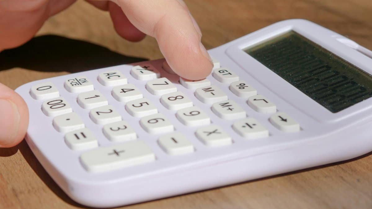 A hand pressing keys on a white calculator placed on a wooden desk in bright daylight.