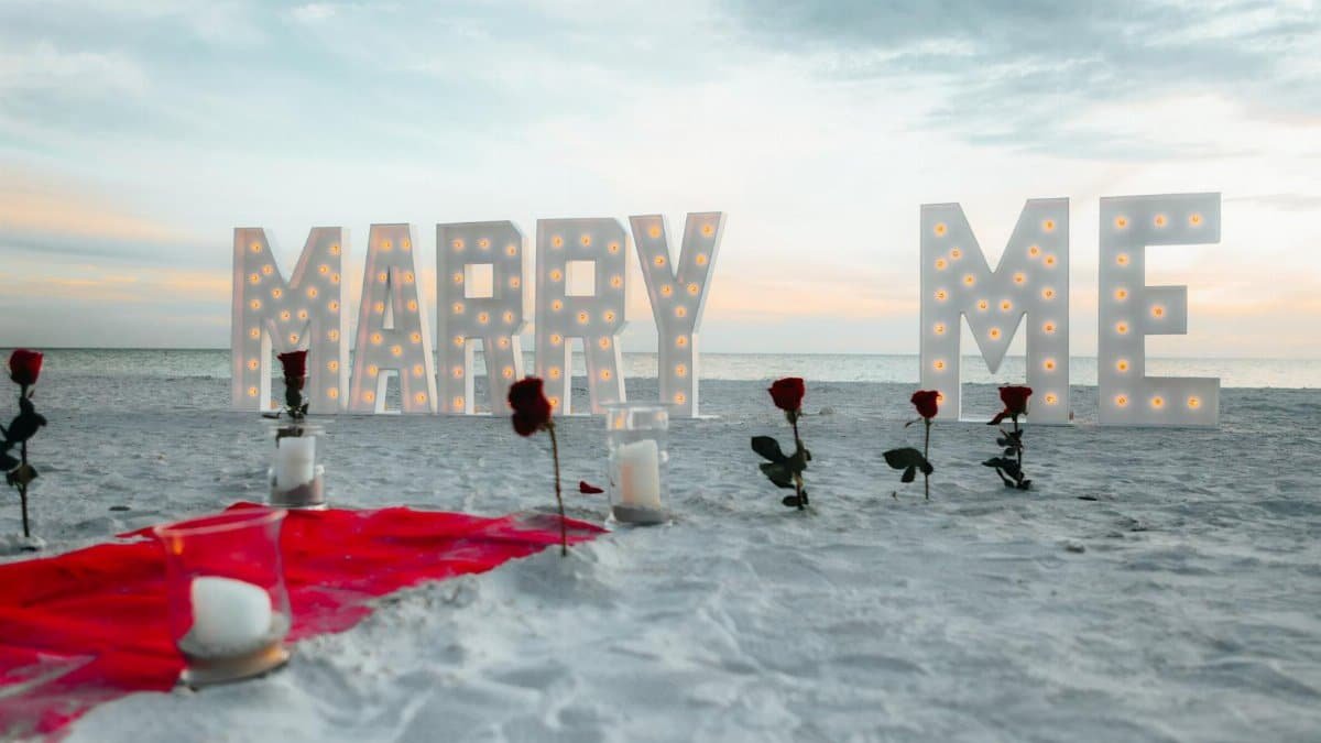 Beach proposal setup with roses and illuminated 'Marry Me' sign at sunset.