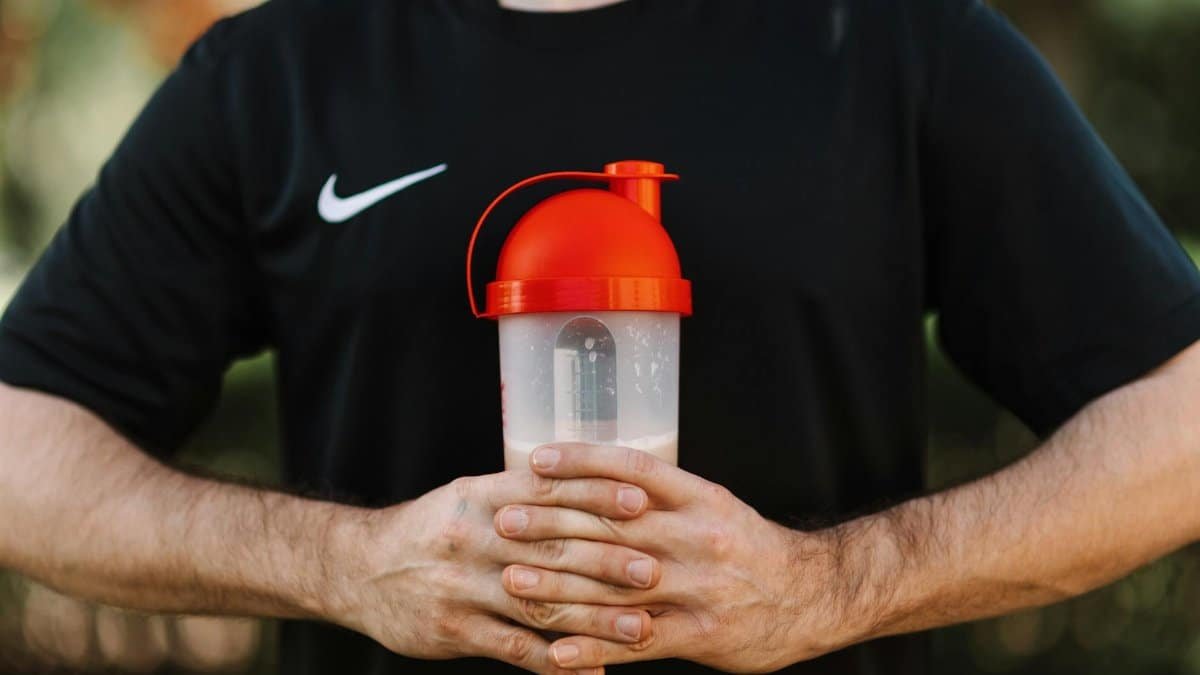 Close-up of a man holding a protein shaker outdoors, wearing a black shirt.