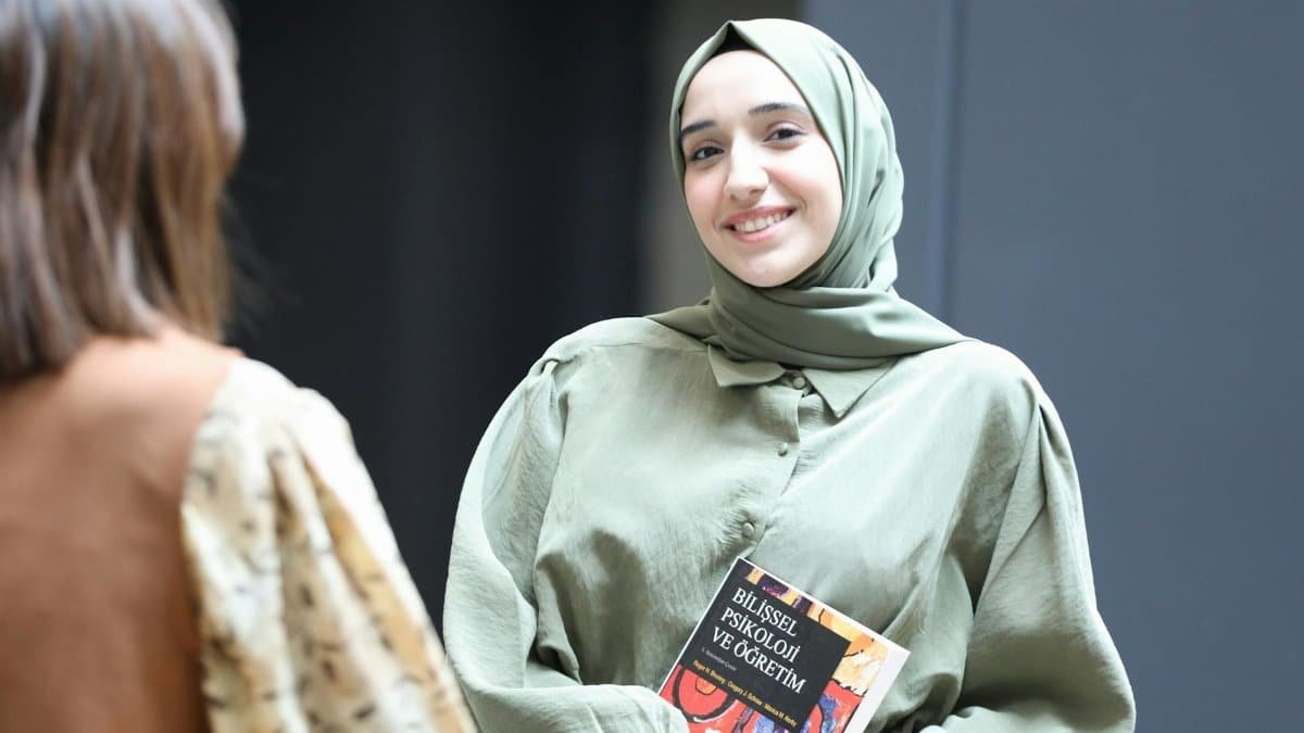 A young woman in a headscarf smiles while holding a book, indoors.