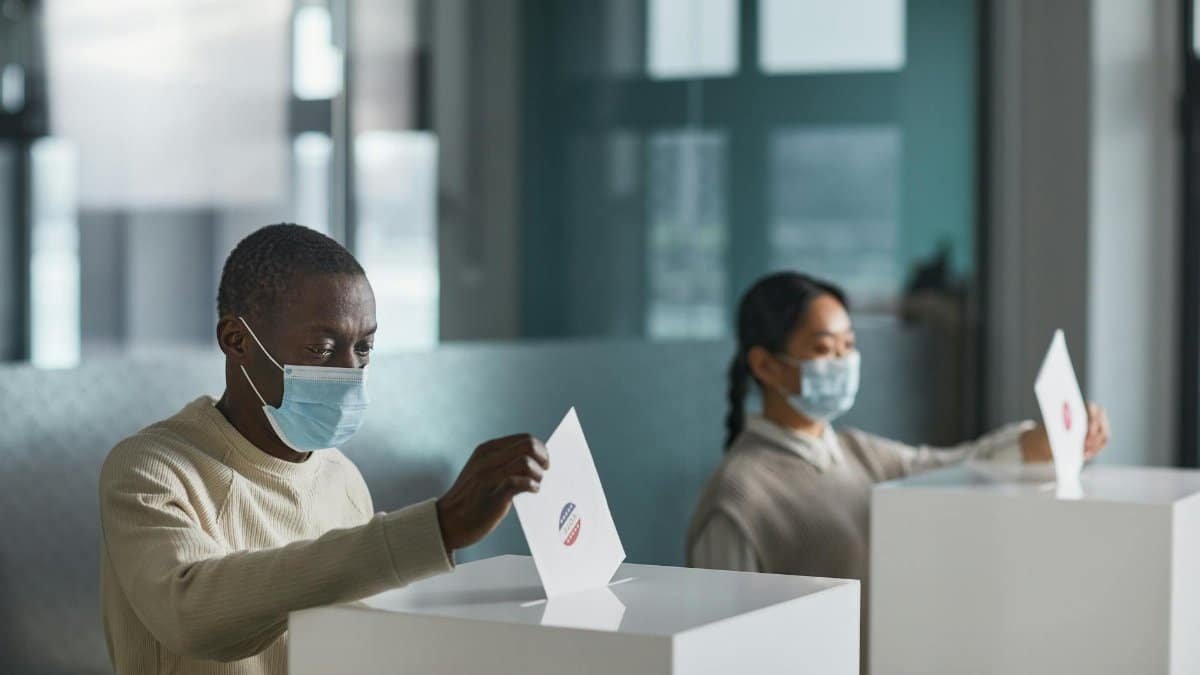 People wearing face masks cast ballots in a public indoor polling station with social distancing.