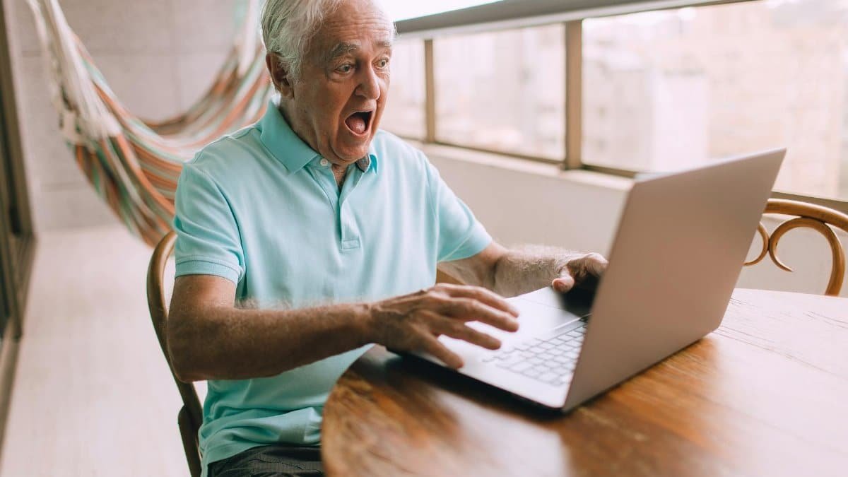 Elderly man in a hammock chair surprised while using laptop indoors. Bright and candid moment.