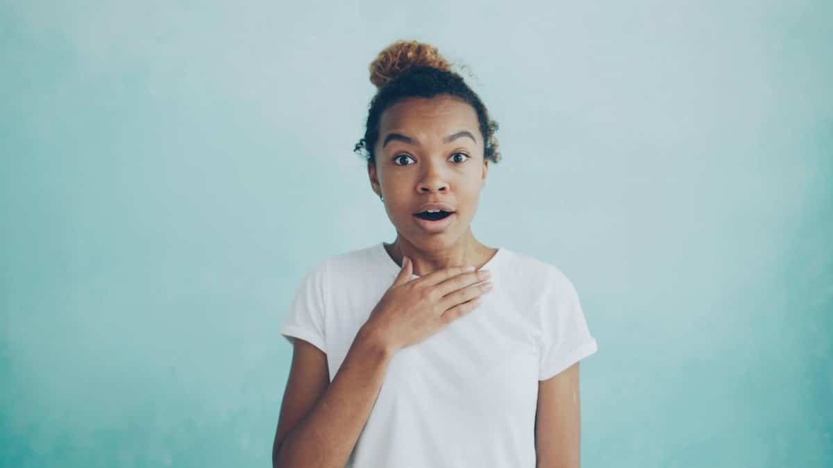 A woman expresses surprise against a soft blue background, wearing a white shirt.