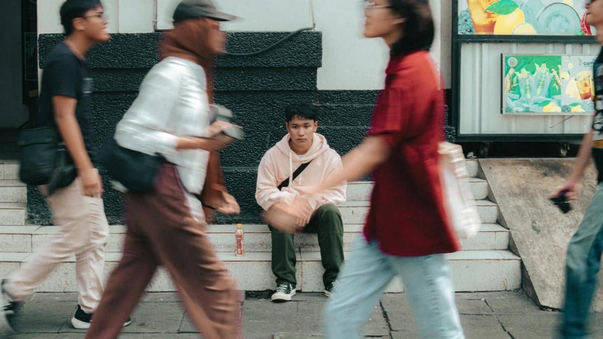 A young man in a pink hoodie sitting on steps as people walk by in motion blur.