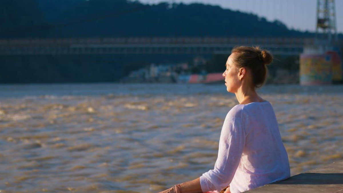 Woman meditating near Ganges River in Rishikesh, India with a bridge in the background.