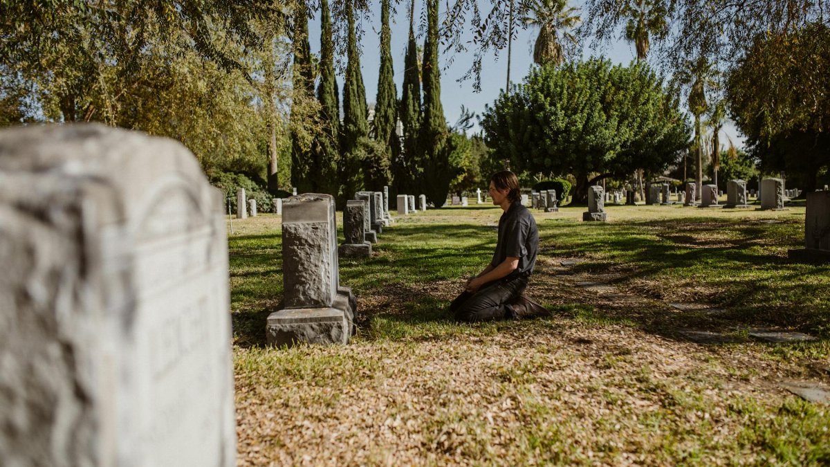 A man kneels in a serene cemetery, reflecting on life and loss, surrounded by lush greenery.