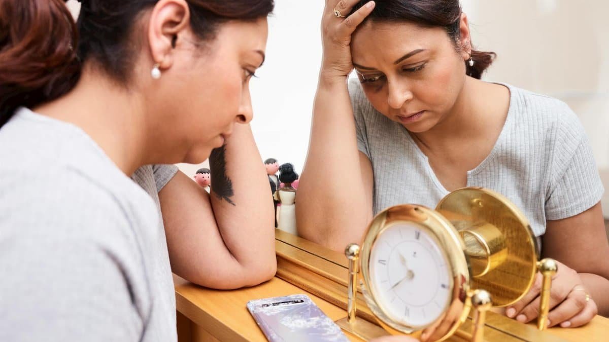 A woman looks pensive and tired, reflecting on burnout with a clock and mirror.