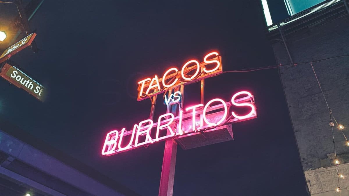 Colorful neon sign displaying 'Tacos vs Burritos' in an urban street setting at night.