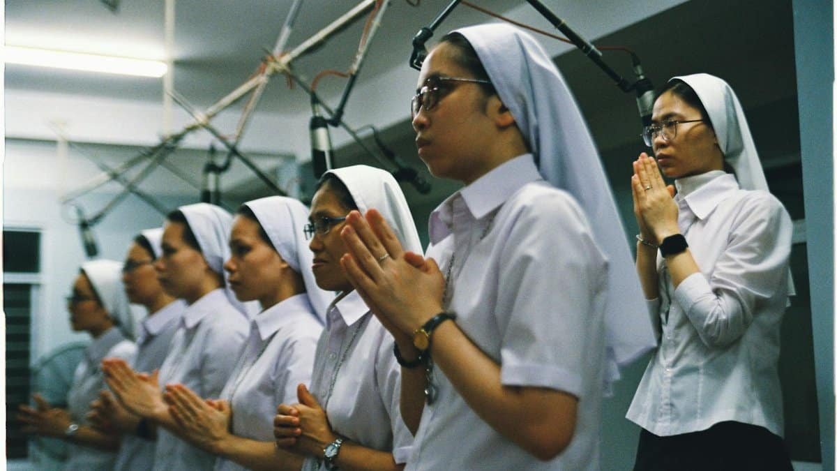A group of nuns in white habits praying during a religious ceremony indoors.
