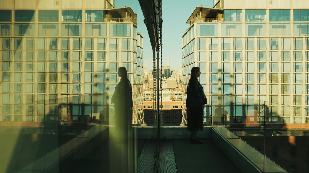 A woman stands on a New York balcony with reflections on a glass facade, showcasing an urban skyline.