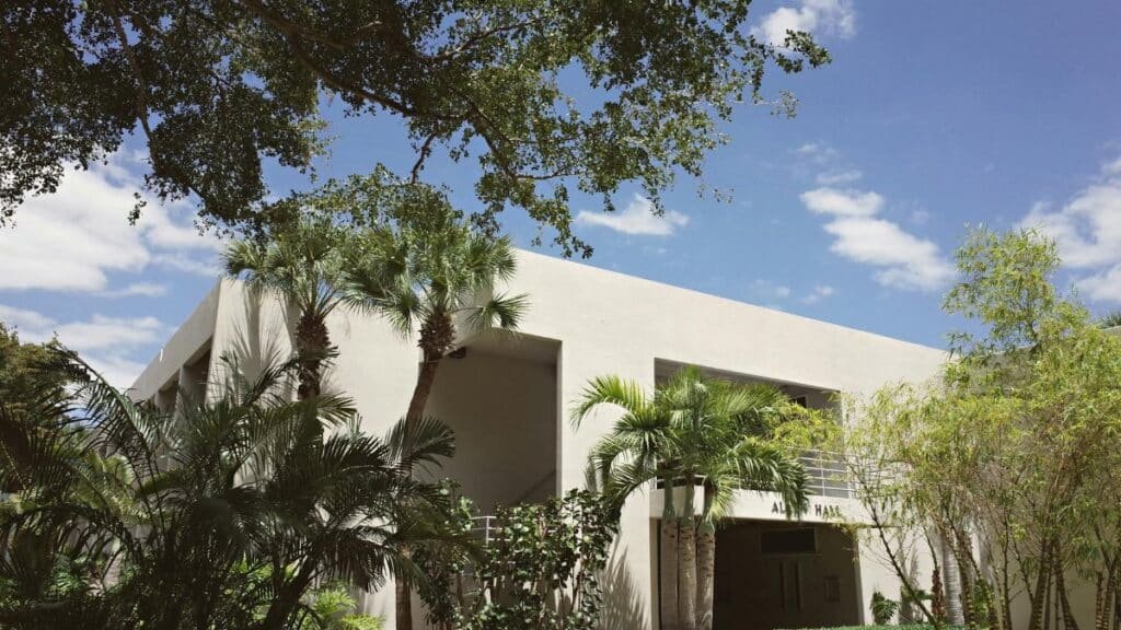A modern university building in Coral Gables, Florida, surrounded by lush palm trees, on a bright sunny day.