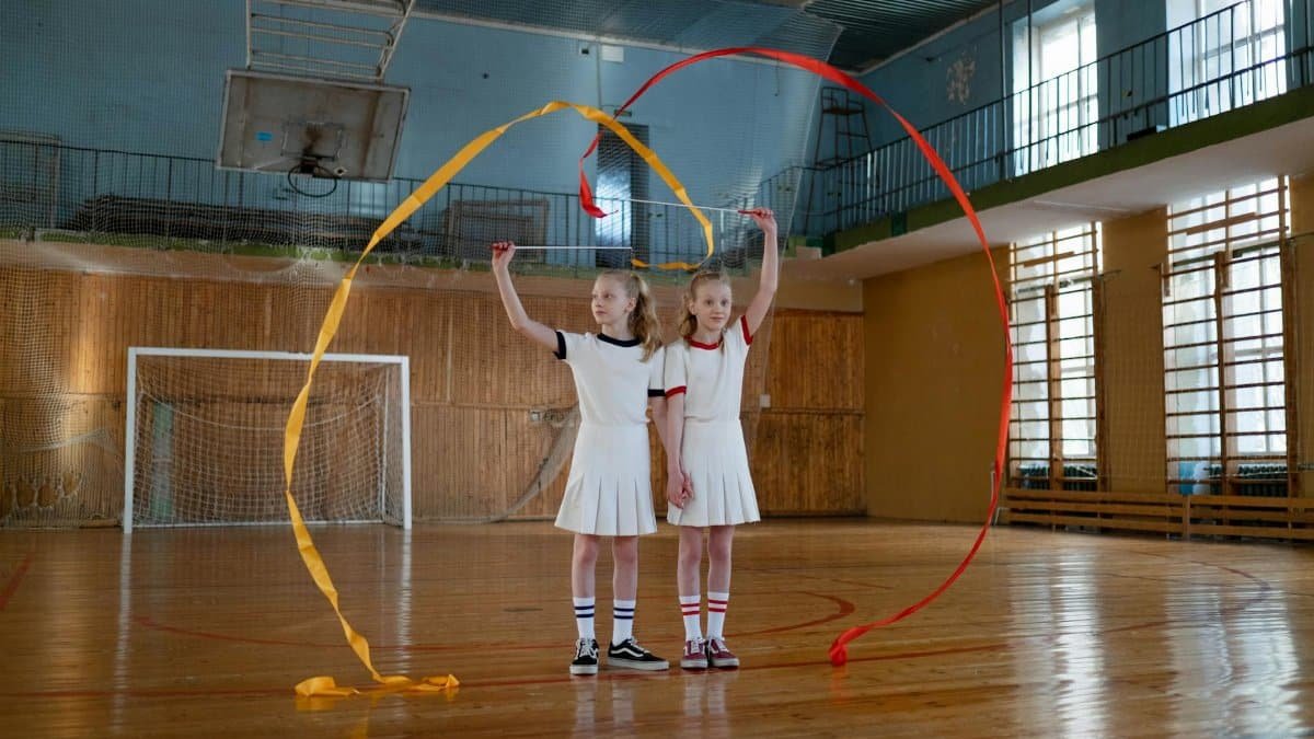Two young girls performing rhythmic gymnastics with ribbons in an indoor gym.