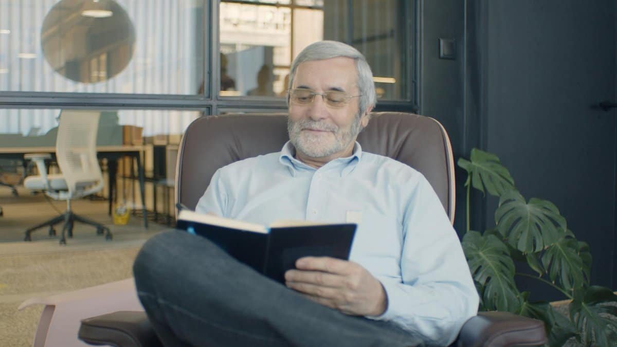 A senior adult with a beard and eyeglasses reading a book comfortably in a modern office setting.