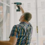 Back view of worker in checkered shirt drilling window frame during renovation process in apartment