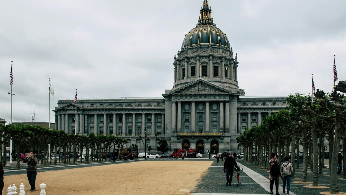 A view of San Francisco City Hall with a public square, trees, and people under a cloudy sky.