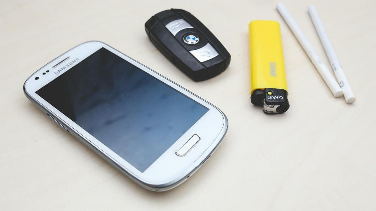 Flat lay of everyday items: smartphone, car key, lighter, and cigarettes on a table.