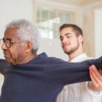 A senior man is assisted by a caregiver for stretching exercises indoors.