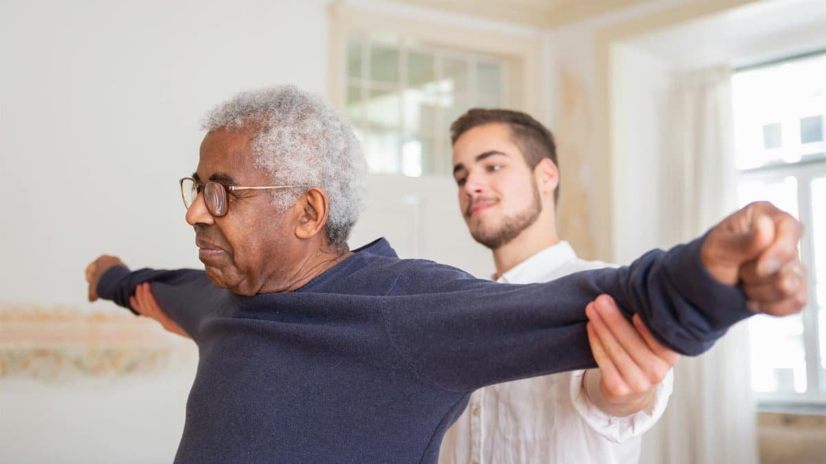 A senior man is assisted by a caregiver for stretching exercises indoors.