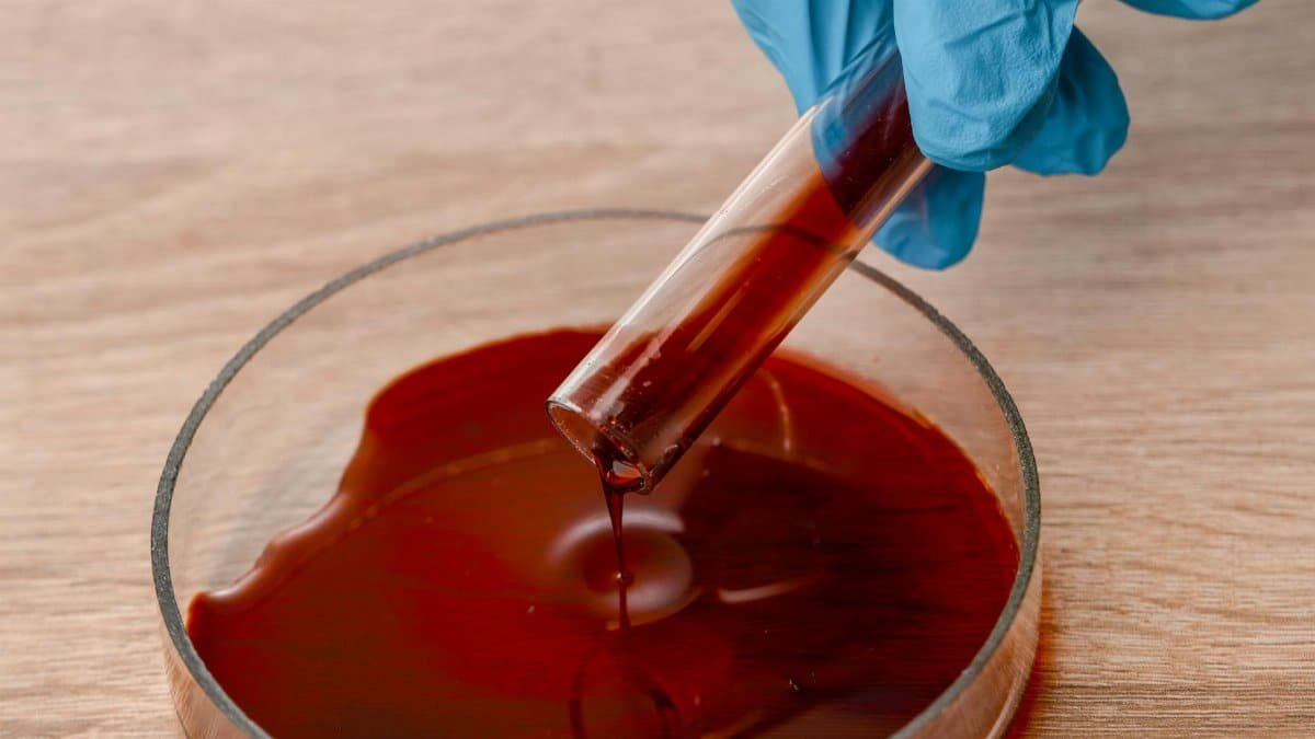 Close-up of gloved hand pouring red liquid from a test tube into a petri dish during a scientific experiment.