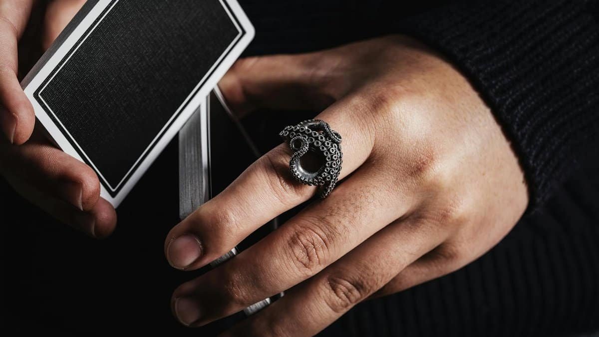 Detailed close-up of a hand wearing an octopus tentacle ring holding playing cards. Dramatic lighting accentuates intricate jewelry design.