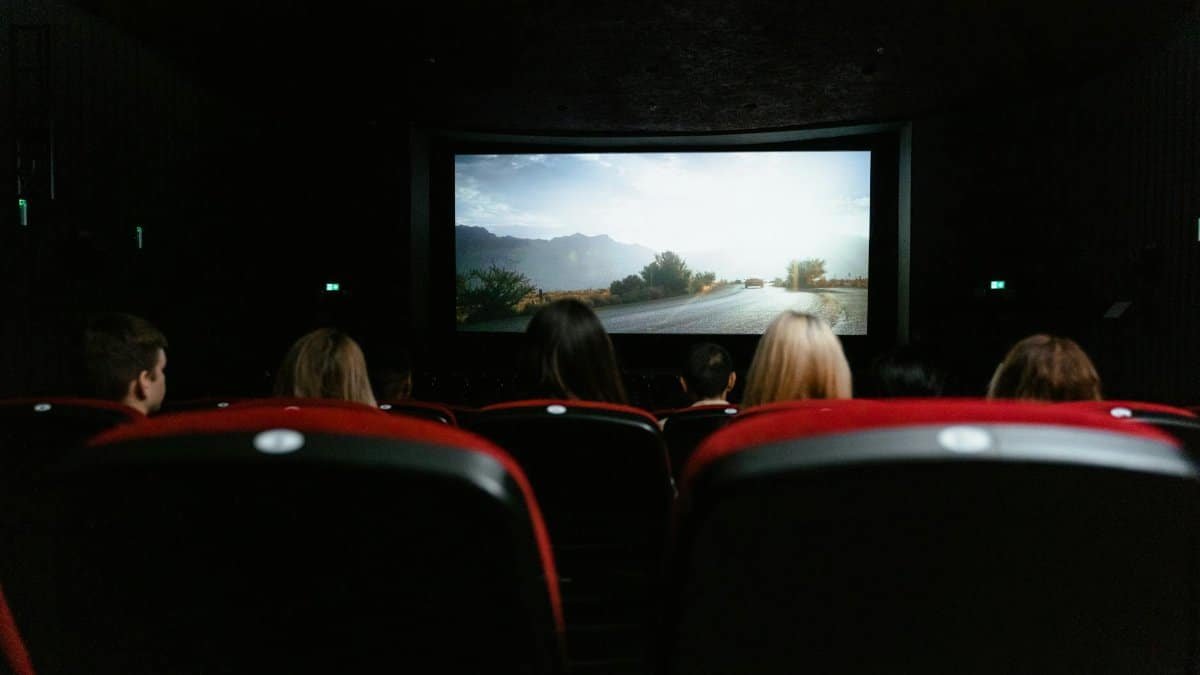 View from behind of people watching a movie in a cinema with red seats and a large screen.